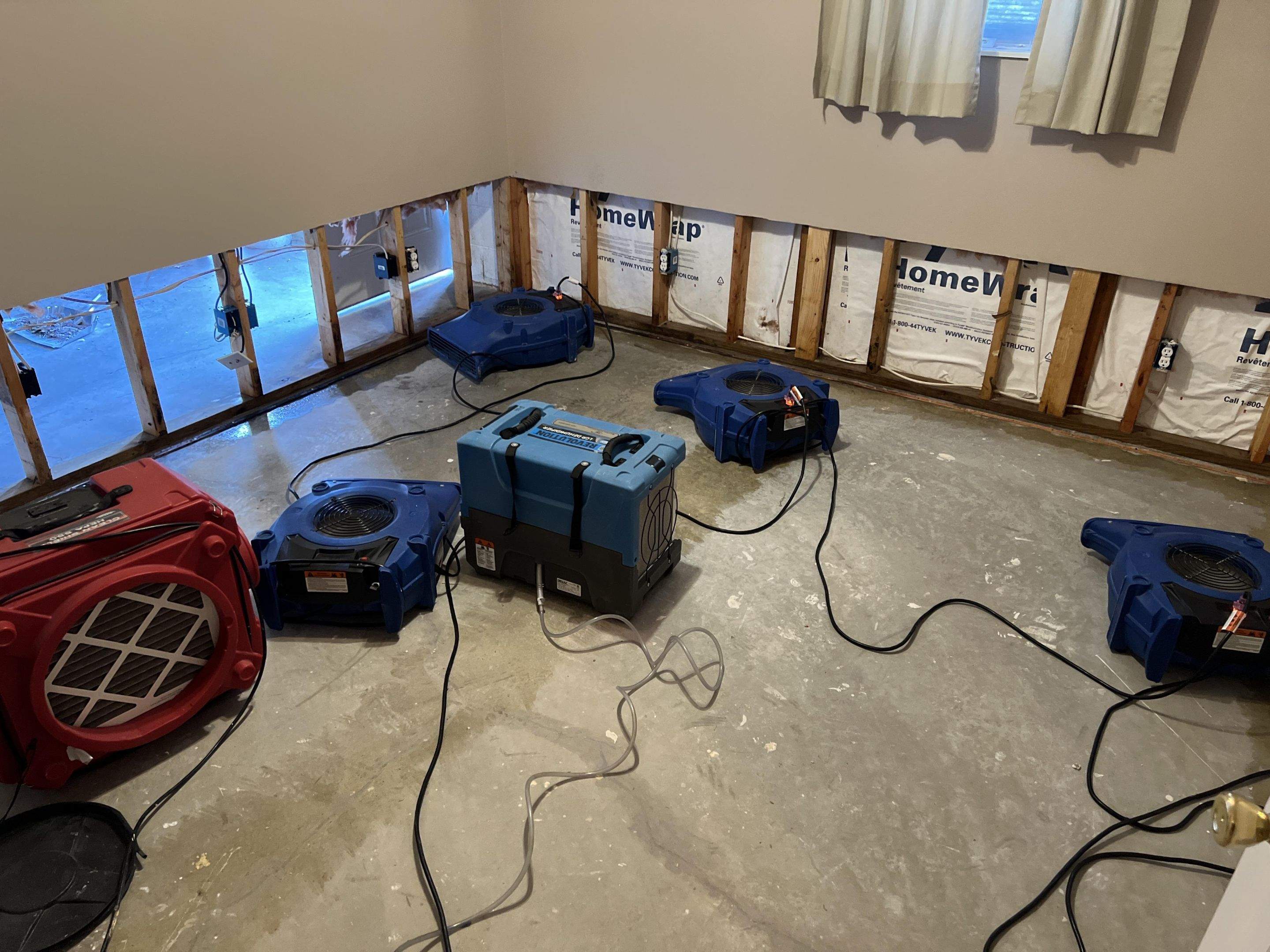Air movers and a dehumidifier drying a room with exposed lower wall framing after indoor water damage